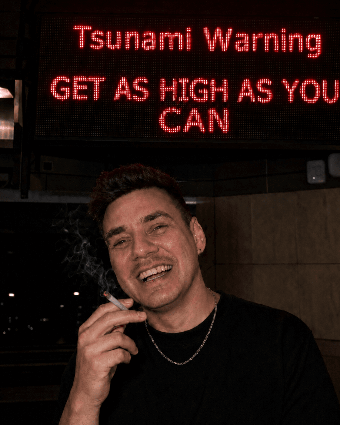 A man smoking and grinning at the camera under a red LED sign that reads "Tsunami Warning — GET AS HIGH AS YOU CAN."