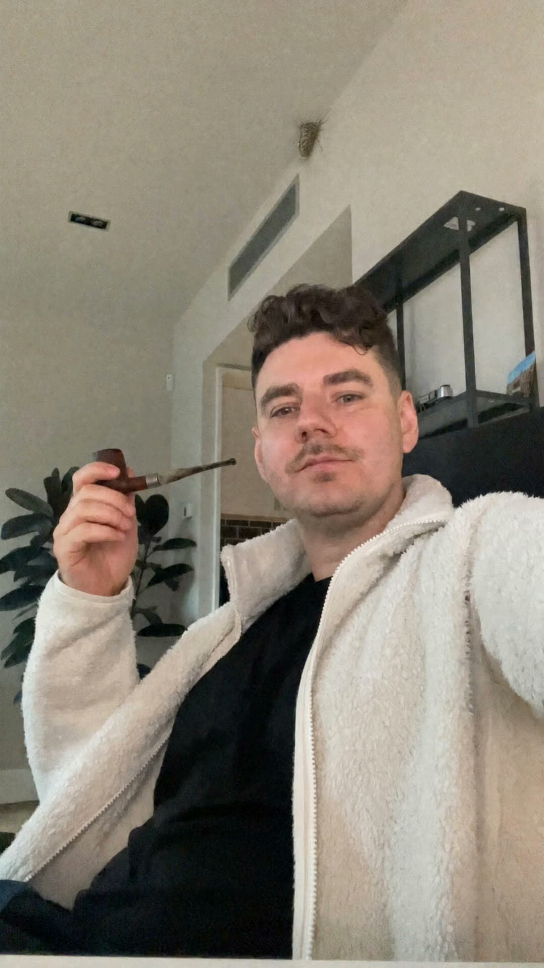 A man in a cream sherpa jacket and dark shirt sits in a sunlit living room, holding a wooden pipe with white smoke curling from the bowl; a small cocoon-like form clings to the high ceiling above him.